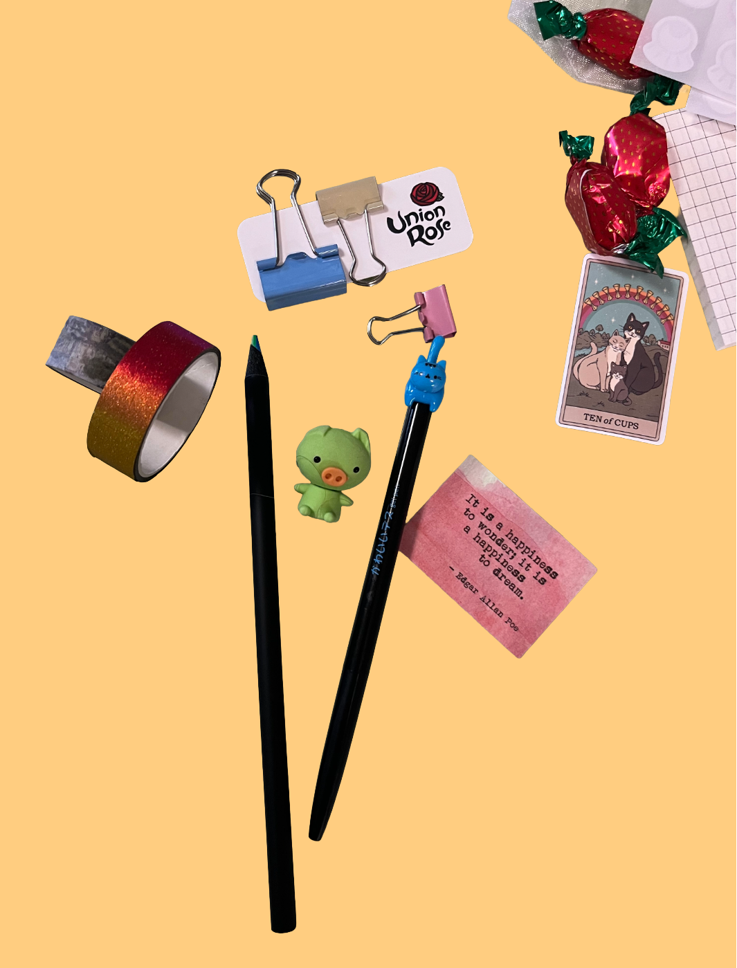 A styled flat-lay of various stationery and small accessories on a solid pale yellow background, including two black pencils, a roll of metallic tape, binder clips, a small green character figure, a pink note, and wrapped red candies.