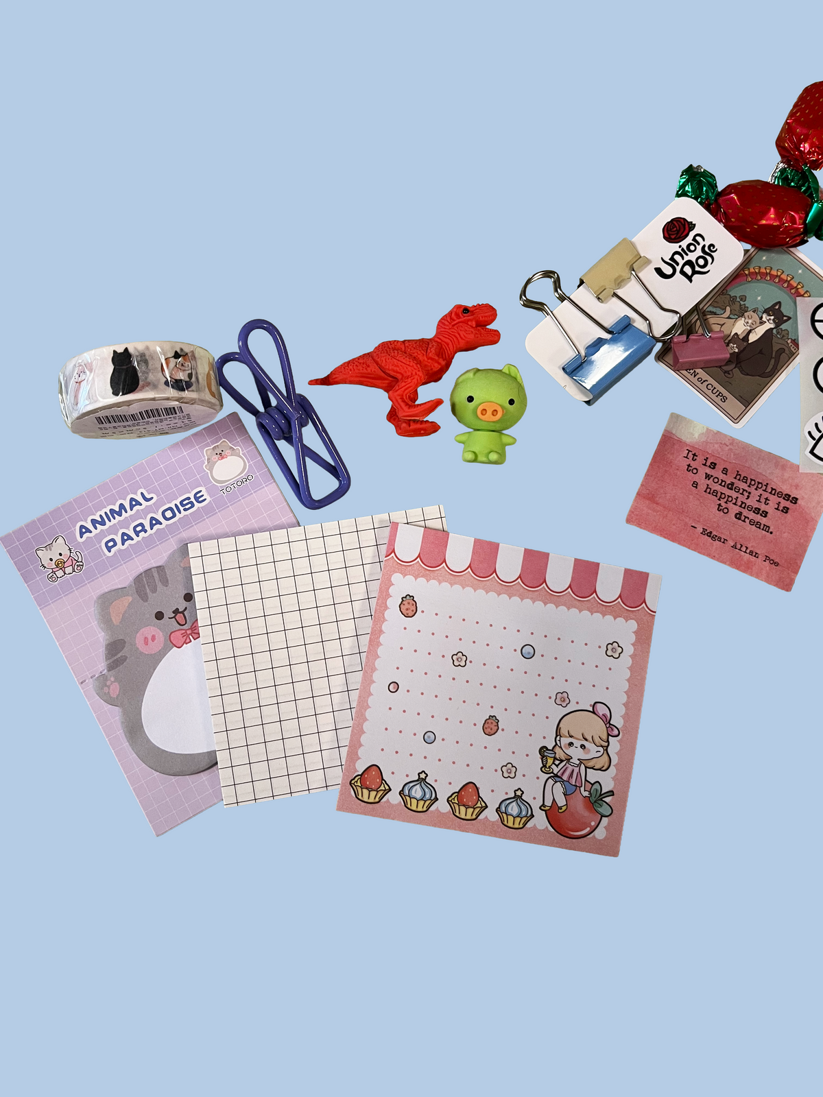 A flat-lay arrangement of cute stationery and small accessories on a light blue background, including a pink 'Animal Paradise' notepad, a grid notepad, a pink cupcake notepad, a purple paperclip, a red dinosaur toy, a green pig toy, binder clips, and candy.