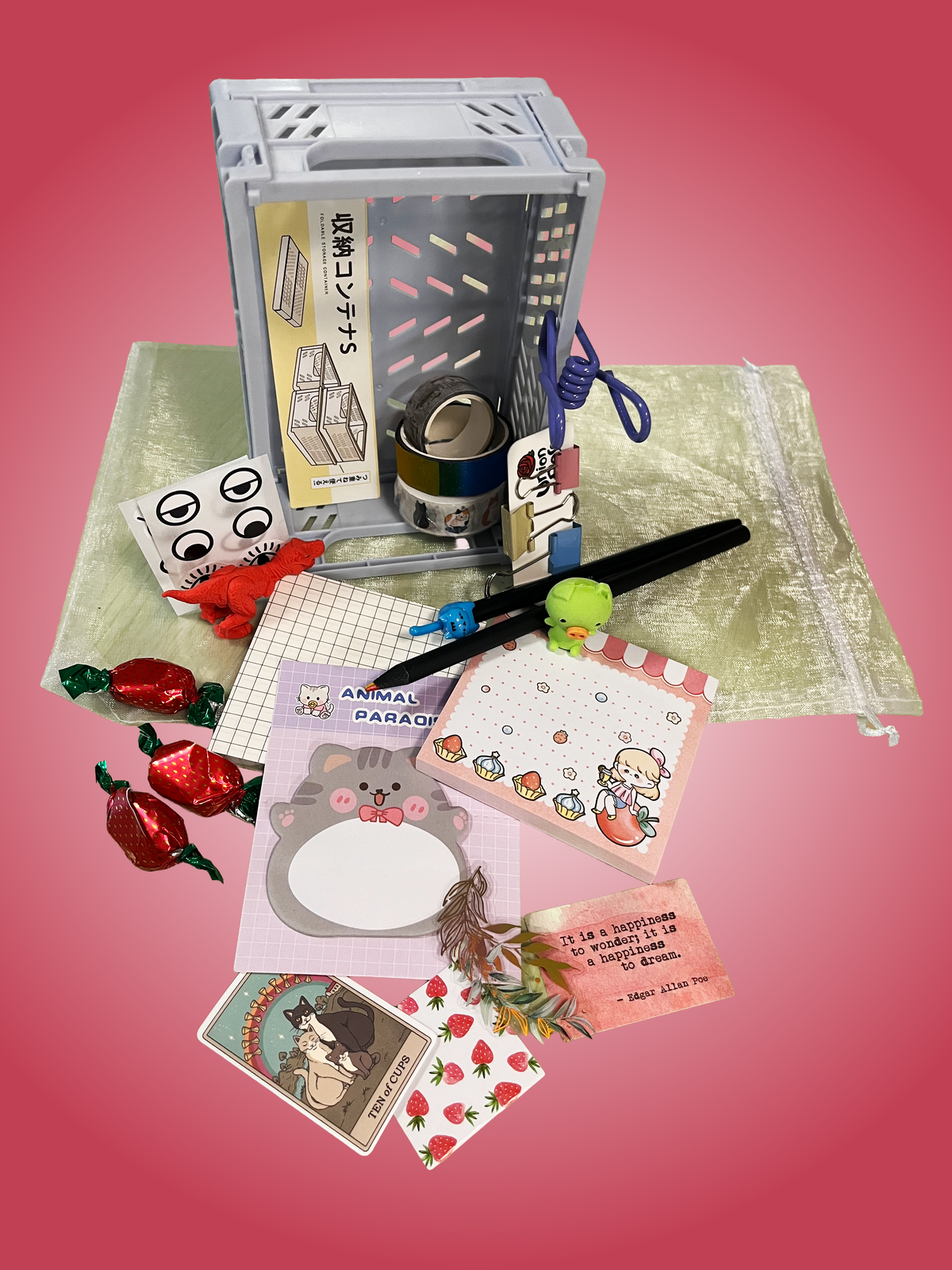 A styled flat-lay of a cute stationery and accessory set arranged on a pale green drawstring bag against a pink gradient background. The set includes a gray collapsible storage box, a patterned notepad, a bear-face memo pad, a small plush green character, a black pen, a blue cord, candy-like red wrapped pieces, a sticker sheet, and several printed cards.