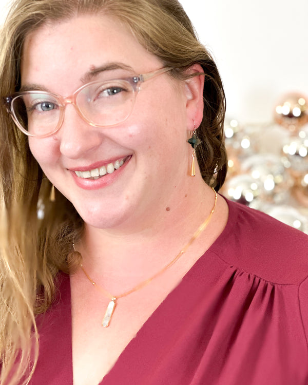 A white woman in glasses modeling a pair of earrings with a green stone anedgold drop. 